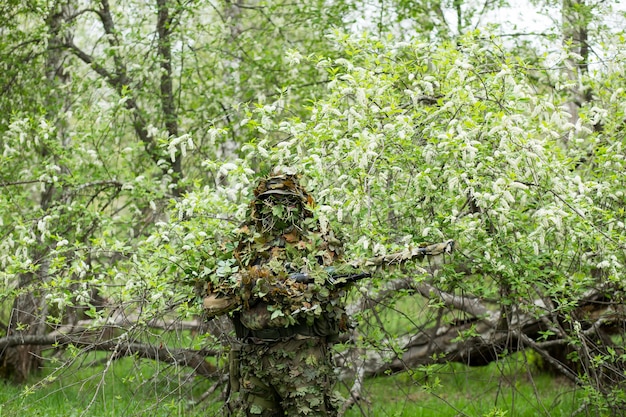 Atirador masculino posando com roupa de camuflagem de camuflagem verde com uma arma, um rifle na mão na floresta perto das flores de cerejeira. Exército, militar, airsoft, conceito de passatempo