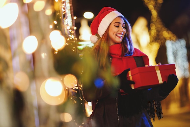Atirar de uma jovem alegre com caixa de presente vermelha está se divertindo na rua da cidade na época do Natal.