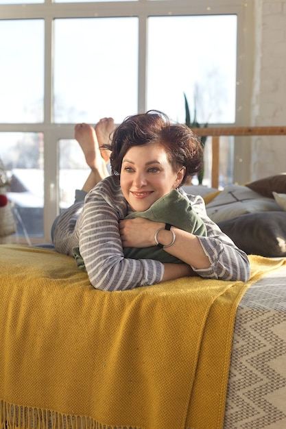 Foto ativa, bela mulher de meia-idade com um lindo sorriso. deitada na cama do quarto abraçando um travesseiro