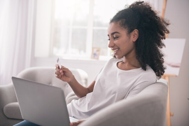 Atividade agradável. Mulher alegre e feliz sentada em frente ao laptop com um cartão de crédito enquanto faz compras online