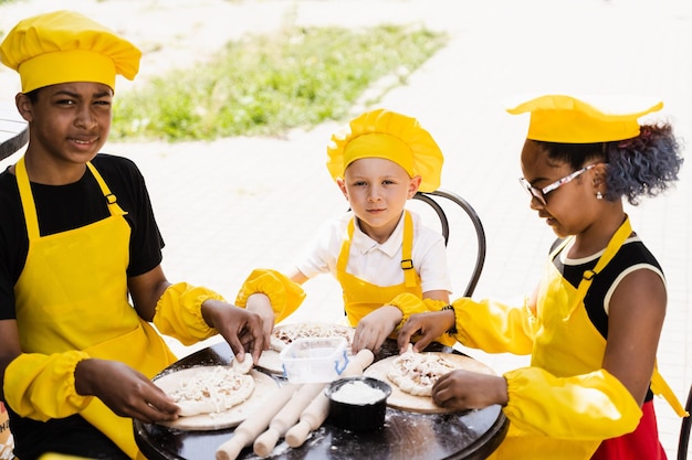 Atividade de cozinhar na infância de crianças multinacionais de crianças negras africanas e caucasianas em chapéu de chef amarelo e uniforme de avental Companhia feliz de crianças multiétnicas cozinhando massa