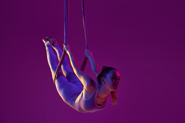Atividade relaxante Jovem garota flexível fazendo treinamento de ioga aérea sobre fundo de estúdio roxo em luz neon Conceito de fitness e estilo de vida saudável