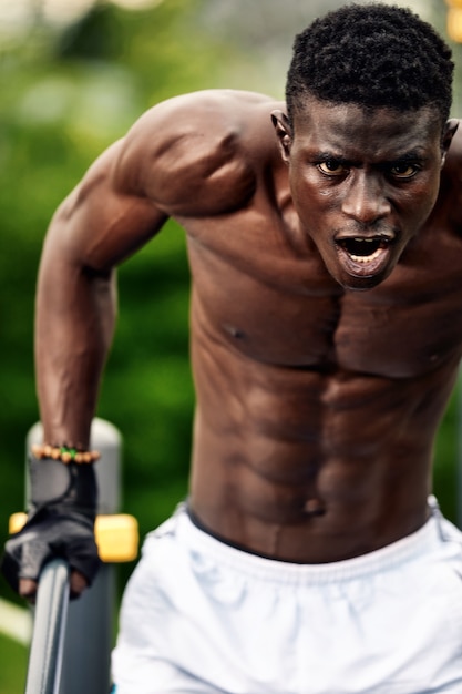 Atleta afroamericano musculoso con un torso esculpido desnudo haciendo flexiones en las barras asimétricas en el paisaje urbano, tiro frontal