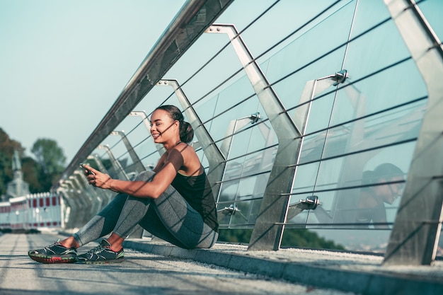 Atleta alegre sentado cerca de la barandilla de vidrio en el puente con el teléfono inteligente en sus manos y sonriendo