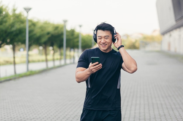 Atleta asiático masculino en una carrera matutina en el parque cerca del estadio escucha música y podcast en grandes auriculares usa el teléfono