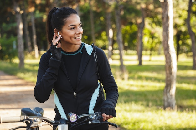 Atleta atraente em forma com uma bicicleta no parque, ouvindo música com fones de ouvido sem fio