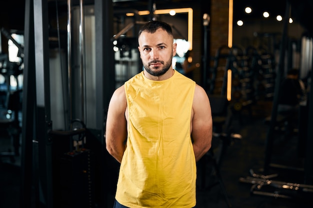 Atleta en camisa amarilla sin mangas de pie en el gimnasio con las manos detrás de la espalda