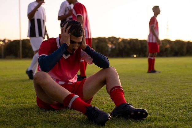 Atleta caucásico triste con la cabeza en las manos sentado en tierra con jugadores en el fondo en el patio de recreo