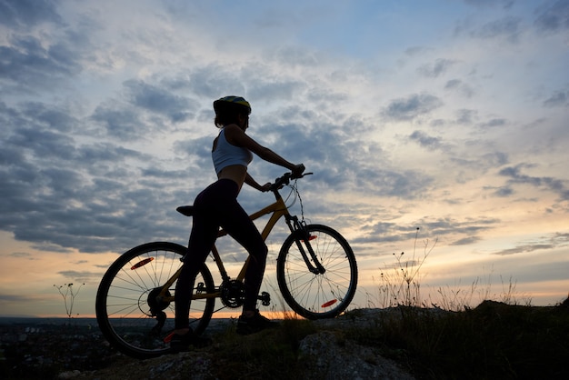 Atleta ciclista feminina com bicicleta na rocha sob o céu da noite linda com nuvens