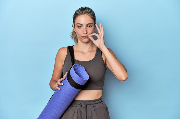 Atleta con una colchoneta en un estudio azul con los dedos en los labios guardando un secreto