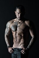 Atleta com músculos bíceps e tríceps homem stripper mostra torso sexy esportista com barriga tanquinho e stripping de ab moda homem com tatuagem tirar jeans desejo e atração em filtro vintage