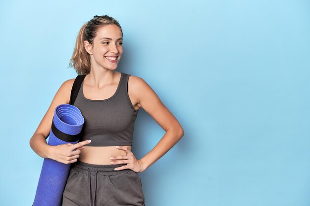 Atleta com um tapete em estúdio azul sorrindo e apontando para um lado mostrando algo em espaço em branco