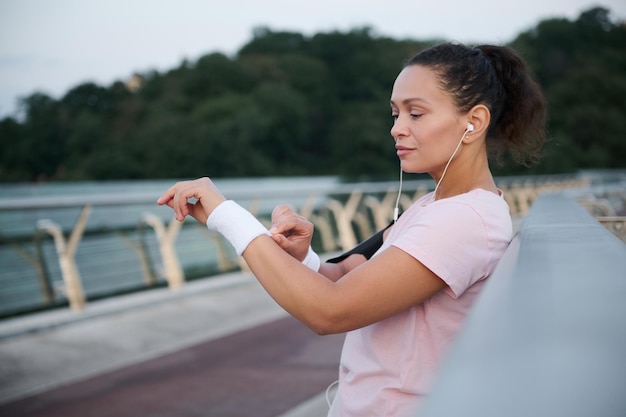 Atleta concentrada em fones de ouvido e uma camiseta rosa está em uma ponte da cidade e usa pulseiras de tênis terry antes da corrida matinal e do treinamento cardiovascular. Mulher bonita atleta malhando ao ar livre