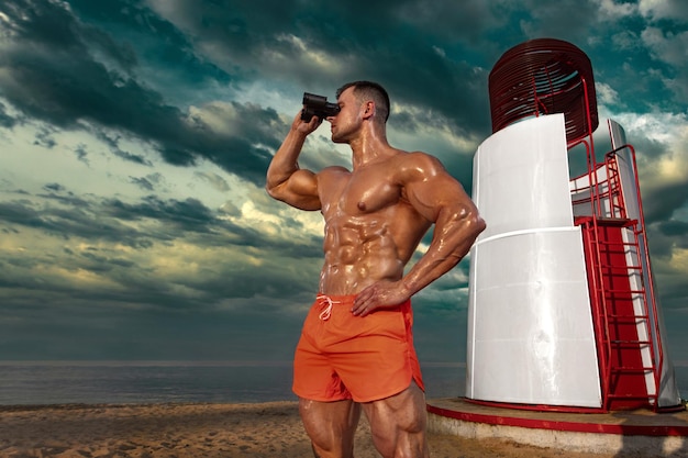 Atleta culturista en forma con binoculares en la mano en la playa Atractivo joven salvavidas cerca de la torre de observación