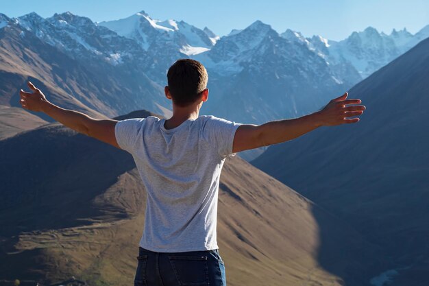 Atleta de camiseta branca em pé no topo de uma colina olhando para grandes picos de montanhas esticando os braços sob a luz do sol.