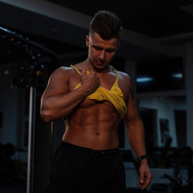 Atleta de cara musculoso sexy com um corpo bronzeado levantou uma camiseta e mostra os músculos abdominais em um fundo escuro. Fisiculturista treinando na academia