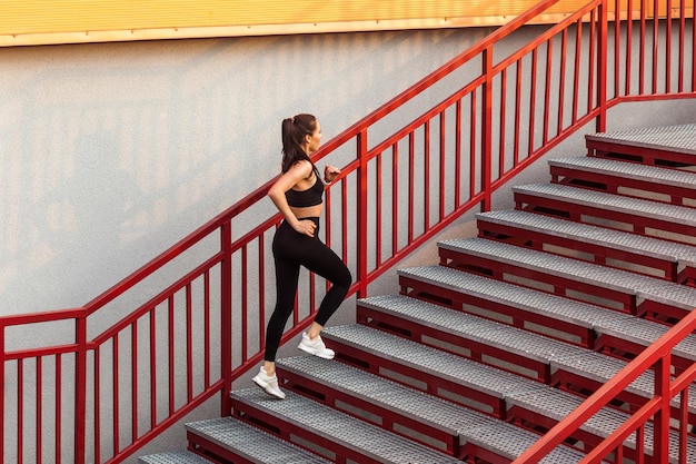 Atleta de corredor cabe menina morena em calças pretas esportivas apertadas e top subindo escadas fazendo exercícios de corrida de treinamento cardio para perda de peso Conceito de cuidados de saúde atividade esportiva ao ar livre