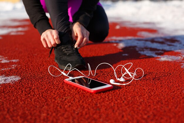 Atleta de garota de tênis preto, agachado na pista vermelha para correr. próximo é um telefone com fones de ouvido com fio. tempo frio de neve