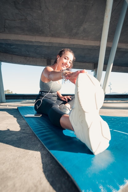 Atleta de linda garota, pele bronzeada africana. Sorri para a câmera enquanto aquece os músculos antes de fazer o treinamento de ioga urbana. Articulações das pernas ginástica articular. Música durante o treinamento. Leggings, roupas e top.