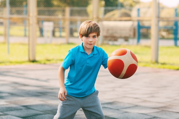 Atleta de menino bonito focado conduz a bola em um jogo de basquete. Um menino joga basquete depois da escola