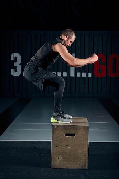 El atleta dio ejercicio. Saltando sobre la caja. Fase de aterrizaje. Fotos de estudio en tono oscuro. El atleta está realizando saltos de caja