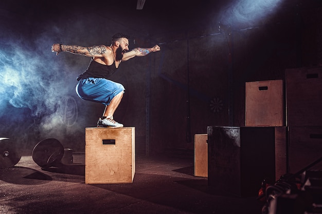 El atleta dio ejercicio. Saltando sobre la caja. Fase de aterrizaje. Fotos de gimnasio en el tono oscuro. Fumar en el gimnasio.