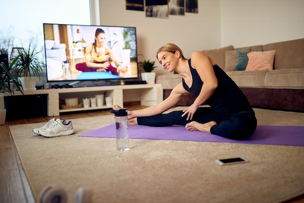 Atleta feliz se esticando no chão enquanto segue as instruções de um instrutor de fitness em uma TV