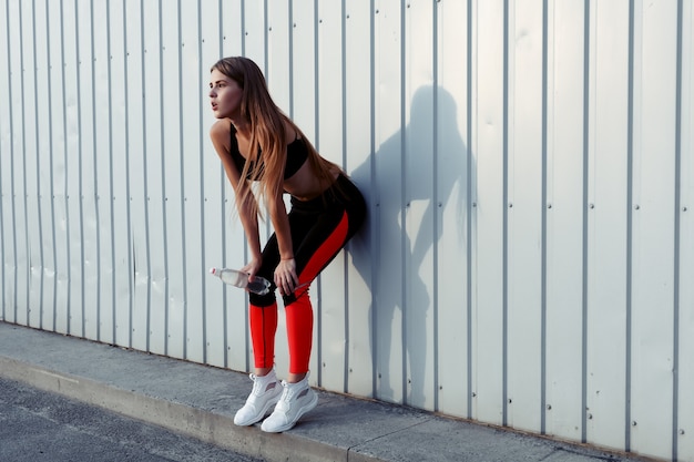 Atleta femenina bebiendo agua mientras está de pie junto a una pared gris. Mujer delgada en ropa deportiva tomando un descanso después del entrenamiento.