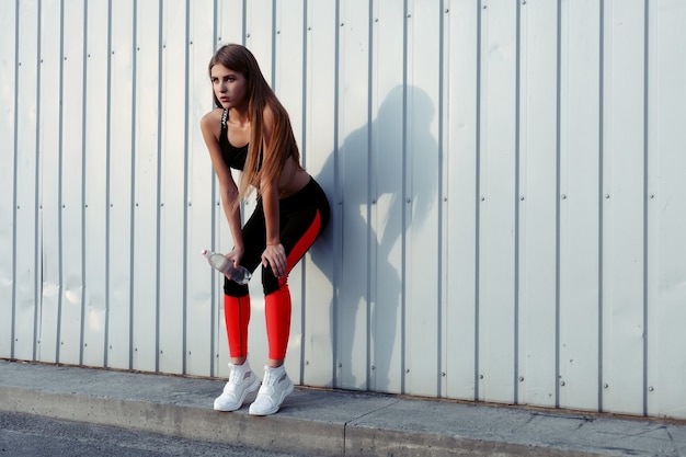 Atleta femenina bebiendo agua mientras está de pie junto a una pared gris. Mujer delgada en ropa deportiva tomando un descanso después del entrenamiento.