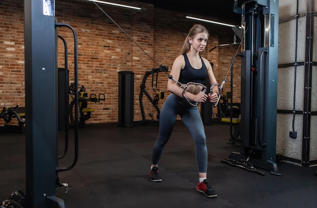 Atleta femenina caucásica joven hermosa que ejercita con la máquina del cable cruzado en el gimnasio.