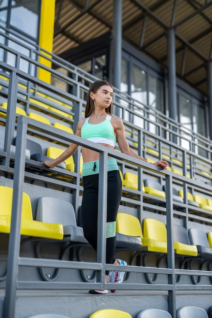 Atleta femenina delgada joven que descansa después del entrenamiento en ropa deportiva de pie cerca del color se sienta en el estadio