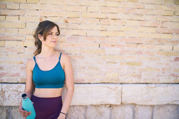 Atleta femenina descansa apoyado contra una pared de ladrillos sosteniendo una botella de agua concepto de estilo de vida saludable