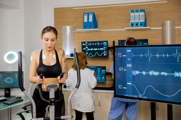 Atleta femenina que hace un esfuerzo físico en el stepper y tiene electrodos adheridos y los pulsos de su corazón se pueden ver en la pantalla. Laboratorio deportivo moderno