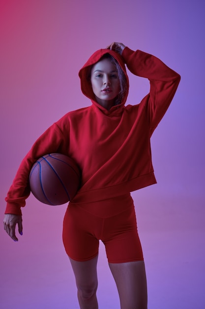 Atleta femenina sexy en sudadera con capucha roja posa con pelota en estudio, fondo de neón