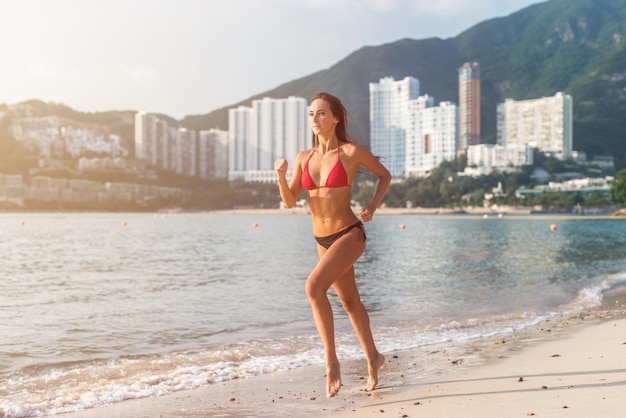 Atleta feminina apta usando biquíni correndo na praia com sol brilhando na câmera e colinas do hotel resort ao fundo