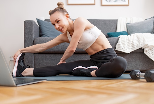 Atleta feminina em forma, flexionando-se na perna e alongando o corpo perto do laptop durante o treino online perto do sofá em casa