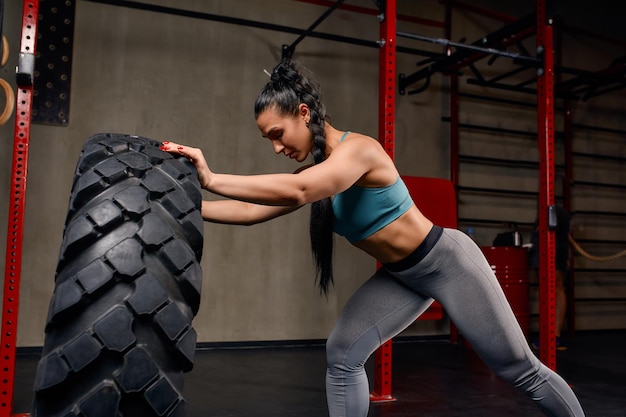 Atleta feminina em forma malhando com um pneu enorme virando e virando na academia mulher exercitando com pneu grande