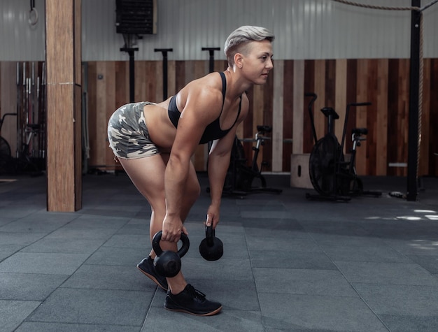 Atleta feminina extraordinária muscular com cabelo rosa curto treina levantamento terra com kettlebells pesados. Funcional, treinamento cruzado no ginásio moderno.