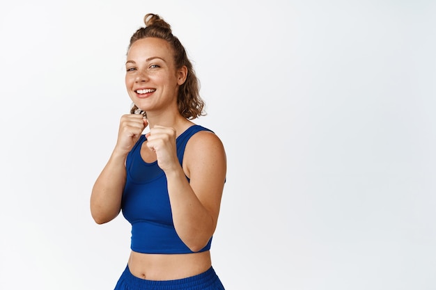 Atleta feminina feliz em pose de lutador, boxe de sombra e rindo, fazendo exercícios de fitness em roupas esportivas, de pé sobre fundo branco