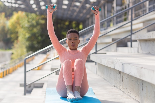 Atleta feminina se exercita com halteres perto do estádio, mulher afro-americana sacode os músculos da imprensa e do assoalho pélvico pela manhã no estádio esportivo