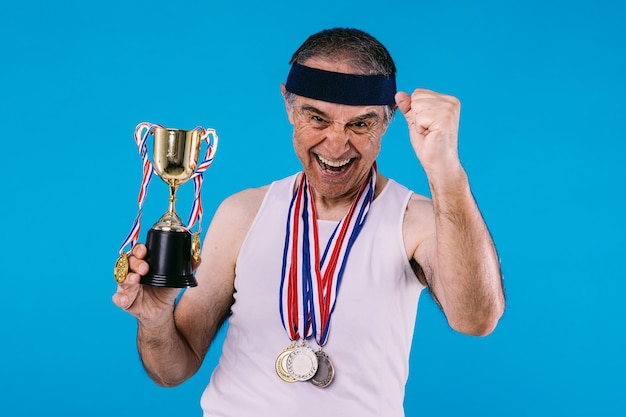 Foto atleta idoso com marcas de sol nos braços, com três medalhas no pescoço e um troféu nas mãos, gritando, sobre fundo azul