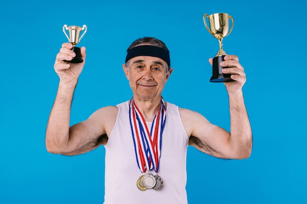 Foto atleta idoso com marcas de sol nos braços, três medalhas no pescoço e dois troféus nas mãos, sobre fundo azul