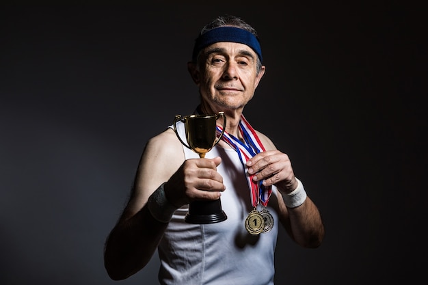 Foto atleta idoso de camisa branca sem mangas, com marcas do sol nos braços, com três medalhas no pescoço, com um troféu nas mãos, sobre fundo escuro. conceito de esportes e vitória.