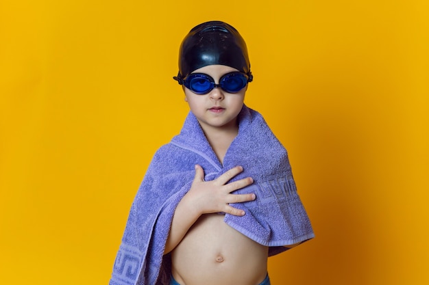 Atleta infantil con gafas de natación azul y una gorra de goma negra y una toalla se encuentra en una pared amarilla