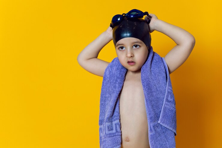 Foto atleta infantil usando óculos de natação azuis e um boné de borracha preto e toalha fica em uma parede amarela
