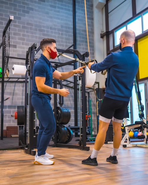 Un atleta con un instructor en el gimnasio haciendo ejercicios de brazos en la pandemia del coronavirus una nueva normalidad Con mascarilla protectora