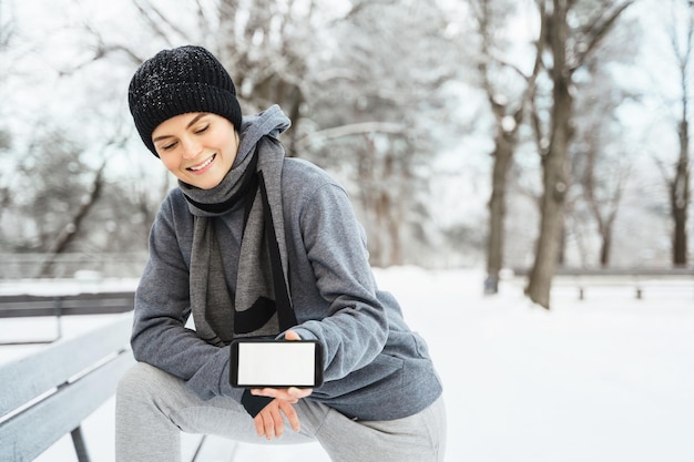 Atleta joven que muestra un teléfono inteligente con una pantalla en blanco durante su entrenamiento de invierno en el parque de la ciudad nevada