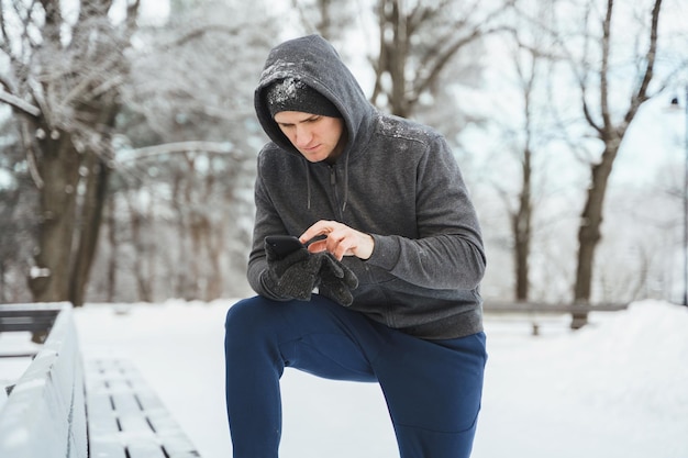 Atleta joven usando teléfono inteligente durante el entrenamiento de invierno en el parque de la ciudad nevada