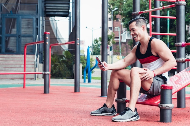 atleta latino joven, atlético, de piel blanca, sentado, hablando por teléfono en un gimnasio, al aire libre.