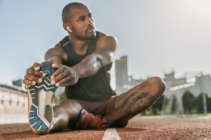 Foto atleta masculino africano musculoso deportivo en auriculares estirando las piernas mientras está sentado en el estadio